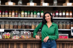  fairytale, 29 year old, straight long black hair, huge tits, extremely cute Mexican woman, (((behind the counter of a little town rustic store))), background of shelves full of food cans,( (two glass shelves one on each side of her,  on top of the counter filled with candies)), she wears a long sleeve green Abercrombie shirt and jeans, she leans towards the counter letting the observer see her nice tits, low intensity lights on the store