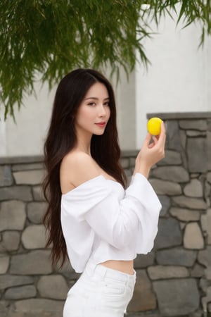 a woman with long dark brown hair, wearing a white off-the-shoulder, long-sleeved shirt and a pair of white pants, is stunning. She is holding a small, round, yellow object in her hands, adding a pop of color to her outfit. The backdrop, a stone wall, and a tree with green leaves, adds a touch of greenery to the scene. A white building can be seen in the background.