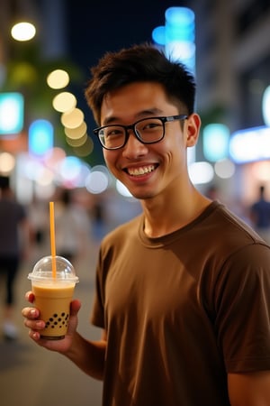 In the photo, an Asian man of medium build with short dark hair, wearing a brown T-shirt, holds a bubble milk tea in his left hand.. He is smiling, with a straw in his right hand. The background is blurred, suggesting a busy city street at night. The man's eyes are wide open, and he's wearing a pair of black-rimmed glasses. The clear cup is filled with a light brown liquid and a straw extends from the top, adding a pop of color to the scene.