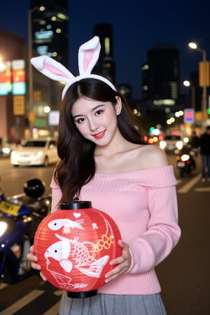 The image is a portrait of a young woman standing in front of a city skyline at night. She is wearing a pink off-the-shoulder sweater and a gray pleated skirt. Her long dark hair is styled in loose waves with white bunny ears and she is looking directly at the camera with a slight smile on her face. The background is blurred, but it appears to be a busy street with cars and motorcycles. The overall mood of the image is casual and relaxed. In her hands, she holds a red and white lantern featuring a large, detailed fish design, adding a festive touch to the scene.

