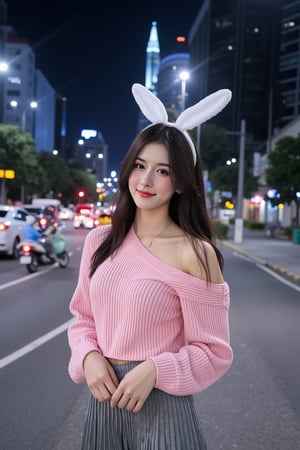 The image is a portrait of a young woman standing in front of a city skyline at night. She is wearing a pink off-the-shoulder sweater and a gray pleated skirt. Her long dark hair is styled in loose waves with white bunny ears and she is looking directly at the camera with a slight smile on her face. The background is blurred, but it appears to be a busy street with cars and motorcycles. The overall mood of the image is casual and relaxed.
