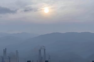 a dense layer of gray clouds illuminated in light rose color from above, a small golden sun, light gray mountains in the distance on the horizon among which power plants and industrial buildings can be seen, closer gray mountains on the horizon among which power plants and industrial buildings can be seen, black mountains on the horizon among which power plants and industrial buildings can be seen,