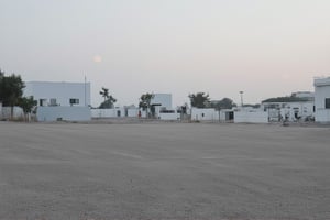 a huge empty asphalt square in the distance one can see massive but not tall rectangular white buildings with narrow black windows, nearby there are smaller rectangular buildings made of brick with square windows and a concrete canopy on the roof, next to them there are not tall trees, a white rectangular building with white edging and large windows, near the buildings you can also see small brick extensions without windows, just in the distance and as if in miniature you can see rectangular buildings with wide large rectangular black windows and white buildings lower but wide with very small black square windows, next to different rare trees are visible behind the buildings, behind all the buildings the yellow sun rises, but it does not dazzle and seems to be in a haze, but all this is as if in the distance and creates the opportunity to examine it all as a whole, and the viewer stands in the middle of perfectly smooth asphalt