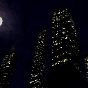 night, dark blue sky with blurry white clouds, full moon, black rectangular skyscrapers with small windows in which white light is visible, metal superstructures are visible at the tops of skyscrapers, brown cubes and a few gray cubes are visible at the base of skyscrapers, the earth is brown mud or clay,