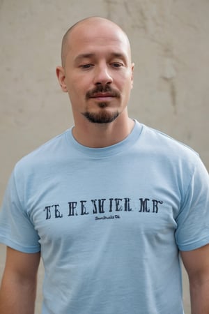 In a medium, eye-level shot, a bald, fair-skinned man with a goatee and mustache gazes downward at the camera, his eyes closed, mouth slightly ajar. He wears a light blue t-shirt with a central blue stripe and the phrase the lord is my shepherd in black letters on the left side, partially visible behind his head. The framing highlights his contemplative expression, with the shirt's design adding a touch of subtle spirituality to the scene.