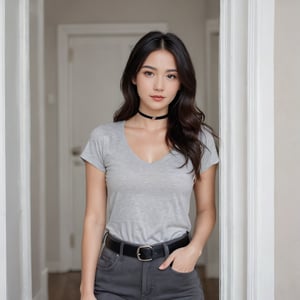 The image is a portrait of a young woman standing in front of a white door. She is wearing a grey t-shirt with a choker neckline and a black belt around her waist. She has long dark hair that is styled in loose waves and is looking directly at the camera with a slight smile on her face. Her left hand is resting on her hip and she is holding a black purse. The background is blurred, but it appears to be a room with a white wall and a door.