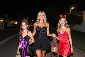 A gorgeous photo of a mother and her two  tween daughters, dressed in sexy Halloween costumes, holding a candy bag, walking on the footpath of a quiet suburban Australian street at night, trick or treaters, Halloween theme, smiling for the camera