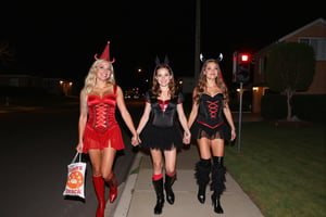 A gorgeous photo of a mother and her two  tween daughters, dressed in sexy Halloween costumes, holding a candy bag, walking on the footpath of a quiet suburban Australian street at night, trick or treaters, Halloween theme, smiling for the camera