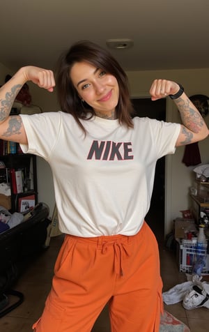 Main frame of a TikTok vertical video featuring a very tattooed punk woman flexing his muscular body toward the camera. She wears a loose oversized "NIKE" t-shirt and loose orange pants, adding a casual, rugged edge to his look. She is in a cluttered bedroom. The composition exudes a laid-back, and rebellious vibe, with a typical adolescent, prideful pose. He gives a mischievous half-smile to the camera, capturing the confident, playful energy.