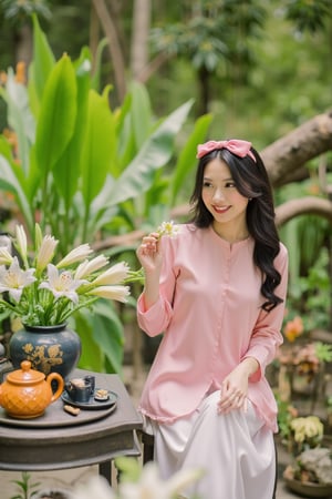 The photo features a young woman seated at an outdoor table in a lush garden setting. She is dressed in a silky pink long-sleeved  top paired with white silk pants. Her long black hair is styled in loose curls, and she wears a matching pink bow headband. The woman is holding a small white flower in her hand, adding a delicate touch to her appearance. On the table next to her, there is a beautiful arrangement of white lilies in a decorative vase, along with an orange teapot and several teacups, suggesting a traditional tea setting. The overall scene is serene and charming, with the vibrant green foliage surrounding the area creating a peaceful atmosphere. The combination of her outfit, the flowers, and the tea set highlights the elegance and cultural richness of the moment, aobaba