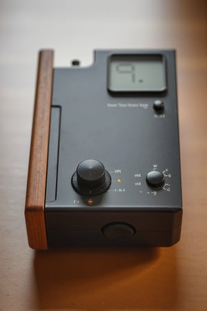 Professional product photo of a device. It features a big button on the front. It also has a screen on the top which shows a measure. The measurement is almost at empty. The device is metallic with wooden accent pieces. It rests on a wooden table. The design follows dieter style. Hasselblad H6D-100c, low exposure, extreme high contrast, ISO 50, with a 100mm macro lens. Light background.