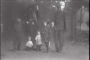 full body shot, a b&w photo from the early 1900's showing an old run down house, a large demonic being, entity about 8 feet tall, long hands, long fingers, with sharp claws, standing on the road next to the house looking scary and meanacing. three young children, in dirty messy clothes, backs to viewer, are standing in front of the demon, waiting for instructions. cloudy day, dark sky, 