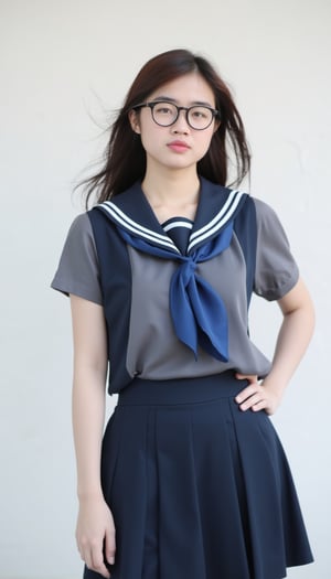 a young woman, dressed in a navy blue and white school uniform, adorned with a blue scarf tied around her neck. She is standing in front of a white wall, her hair cascading over her shoulders, adding a touch of movement to her face. She's wearing a pair of black framed eye glasses, her left hand resting on her hip. Her right hand is draped over her right shoulder, adding depth to the composition.