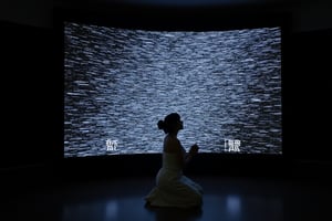 A woman kneels in deep prayer, her hands gently clasped, facing a colossal television screen that towers above her. The TV's screen flickers with chaotic static, a wash of black and white noise that pulses in waves, casting a cold, shifting light over the scene. The woman is bathed in this erratic glow, her silhouette softly illuminated by the constantly shifting patterns of the static. Her figure is serene, dressed in simple, flowing garments that ripple slightly, reflecting the artificial light.

The room around her is stark and minimalist, with the gigantic TV as the focal point. The screen itself is a monolithic presence, slightly curved, and its glossy frame gleams faintly in the low ambient light. The static, constantly moving, seems alive, with random flashes and bands of light darting across the screen, filling the atmosphere with a sense of both unease and wonder.

The visual style blends realism with an unsettling, surreal tone, evoking themes of dystopian futurism. The lighting is harsh yet intimate, with sharp contrasts between the illuminated woman and the darkened surroundings. The static creates a grainy, almost tactile texture against the smooth surface of the TV, while the woman's posture exudes a sense of quiet devotion, creating a powerful juxtaposition between the spiritual and the technological. The overall composition is cinematic, like a still frame from a futuristic, introspective narrative, where technology looms as a vast, enigmatic presence.
