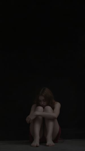 a girl sitting in sorrow, looking sad her head is down, she's wearing a top and skirt, sitting against a wall and the background is Black 