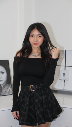 A young Asian woman stands confidently in front of a crisp white wall, dressed in a striking ensemble. Her long, dark hair cascades down her back as she wears a black off-the-shoulder shirt with long sleeves, paired with a plaid black skirt and matching belt featuring a gold buckle. A pop of vibrant red color accentuates her lips, drawing attention to her facial features. Around her neck, a silver chain adorns the necklace that catches the light. To her right, a framed black and white photograph of a woman's face adds an intriguing element to the scene, creating a sense of depth and visual interest.