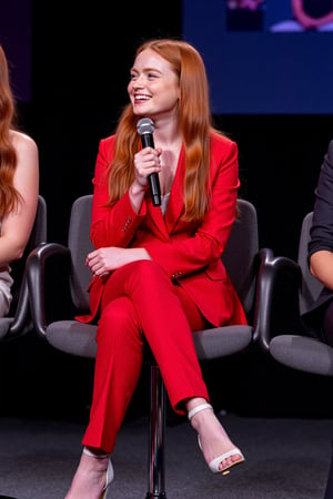 Photography of a redhead girl with long, straight and silky hair, sitting cross-legged on a modern chair, at a press conference on a  open-hair stage. She holds a microphone and looks to her side with a happy expression. She wears a red suit paired with pants and heeled sandals.