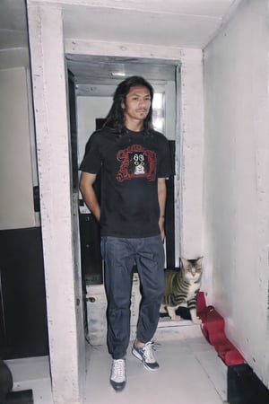 A man, m124n, long tied straight hair with long sideburns, wearing a dark-red colored loose t-shirt, cat cartoon image on the t-shirt, baggy black colored jeans, standing straight and still in a total fully white colored room, flash,M124n,Hair,photorealistic,m124n