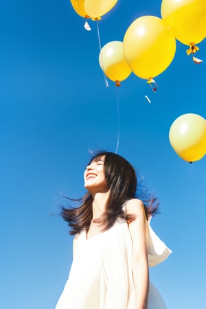 A young Asian woman with short hair, smiling and wearing a white dress, walking in a grassy field during the daytime. Blue skies, white clouds, warm sunlight. A bunch of colorful balloons and flowers floating around her. Wide-angle shot, cheerful and bright atmosphere, natural lighting.