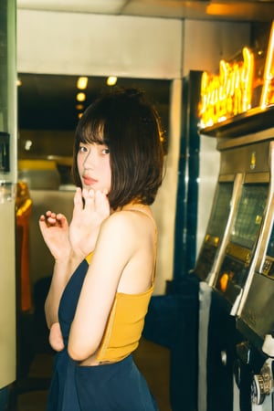 A young Asian woman with short hair, wearing a stylish outfit, squatting in front of a camera in a vintage Japanese laundromat. She is striking a cool pose with a confident look in her eyes, gazing directly at the camera. The scene has a warm yellow tone, evoking a retro atmosphere.