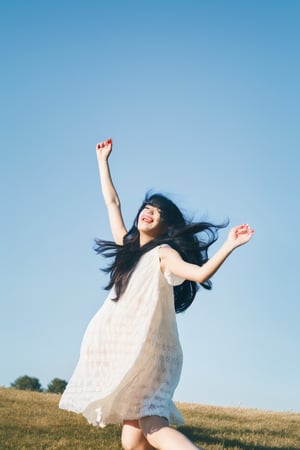 A young Asian woman with short hair, smiling and wearing a white dress, walking in a grassy field during the daytime. Blue skies, white clouds, warm sunlight. A bunch of colorful balloons and flowers floating around her. Wide-angle shot, cheerful and bright atmosphere, natural lighting.