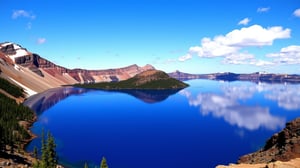 This is a high-resolution photograph capturing a stunning panoramic view of Crater Lake, located in Oregon, USA. The image showcases the deep blue waters of the lake, which reflect the sky and surrounding mountains with remarkable clarity. The lake is framed by the rugged, rocky edges of the caldera, which formed after a volcanic eruption. The caldera walls are a mix of brown and red hues, with patches of green vegetation interspersed. A prominent island, Wizard Island, rises from the center of the lake, covered in lush green trees. The island is surrounded by a vibrant blue water that contrasts sharply with the darker, deeper hues of the lake. In the background, the sky is a bright blue with a few scattered, fluffy white clouds. The photograph captures the serene and majestic beauty of the natural landscape, emphasizing the pristine clarity of the water and the ruggedness of the surrounding terrain. The overall composition is balanced, with the island positioned slightly to the right of the center, drawing the viewer's attention towards it. The photograph is likely taken during the daytime, as the sunlight highlights the textures and colors vividly.