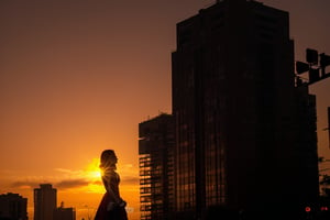 At sunset, the orange-red sky and the silhouettes of high-rise buildings.) (A steady flow of people on the sidewalk.) (A young woman in a red dress stands at the intersection, making a gesture of one hand  holding the sun with her hands through dislocation. Gold The sunlight shines from behind her, giving her silhouette a halo.