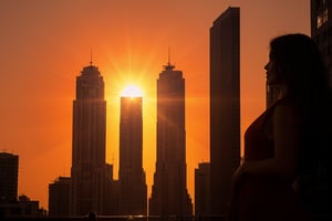 At sunset, the orange-red sky and the silhouettes of high-rise buildings.) (A steady flow of people on the sidewalk.) (A young woman in a red dress stands at the intersection, making a gesture of one hand  holding the sun with her hands through dislocation. Gold The sunlight shines from behind her, giving her silhouette a halo.