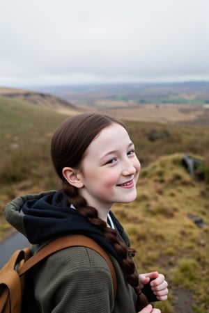 Candid scene of a happy young girl hiking in the hills of Wales. She is tall and slender build, striking features, and long black hair in a long french braid. Her porcelain-like complexion is accentuated by a sprinkling of freckles across the bridge of her nose. She is smiling as she looks out at the scenery. 