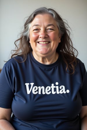 close-up portrait shot of a chubby 80 year old woman wearing a dark blue teeshirt with the word "Venetia."  emblazoned on it in bold white block text, laughing and smiling, gazing into the camera lens at an angle. The subject's long grey hair is wavy and messy, framing her round face against a clean and minimalist background. The overall focus is on the subject's facial features.,