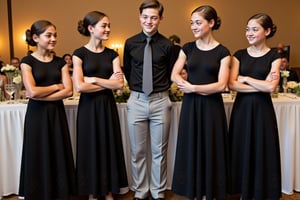 Four teenage girls flank a teenage boy as they casually lean against a tall table at a wedding. Each girl wears a long black cap sleeve dress with black flowers embroidered along the hem. The boy wears a black with a grey shirt and black flower patterned tie. All three have dark tan skin and childish builds. The girl on the left has dark blonde hair and the others have dark brown hair. They are mourning, but happy.