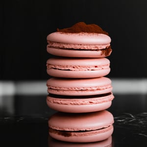 The image showcases a stack of pink macaron,The macaron are arranged in a vertical column, The macaron have a textured surface with a series of ridges with a light dusting of cocoa or powdered sugar on top. The stack is set against a dark, possibly black, background which accentuates the rich colors of the macaron. The lighting is soft and focused on the macaron, creating a moody and appetizing atmosphere. The reflection on the surface beneath the stack suggests a glossy or polished surface, possibly a countertop or a marble slab.