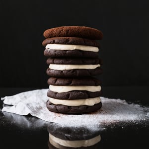 The image showcases a stack of chocolate sandwich cookies, commonly known as Oreos. The cookies are arranged in a vertical column, with each layer consisting of a dark chocolate cookie and a creamy white filling. The cookies have a textured surface with a series of ridges, which is characteristic of the Oreo design. The filling appears to be smooth and creamy, with a light dusting of cocoa or powdered sugar on top, adding a touch of contrast to the dark chocolate. The stack is set against a dark, possibly black, background which accentuates the rich colors of the cookies. The lighting is soft and focused on the cookies, creating a moody and appetizing atmosphere. The reflection on the surface beneath the stack suggests a glossy or polished surface, possibly a countertop or a marble slab.