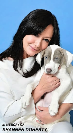 In Memory of Shannon Doherty. A woman with dark hair, wearing a white top, cradles a small puppy in her arms against a blue background. The puppy has a light coat with darker markings on its face and ears.