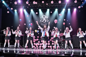 A student in a school uniform stood on the stage holding a guitar and singing with a microphone. The stage was surrounded by five students, three of whom were holding balloons and two of whom- were holding banners. The atmosphere was very lively.


