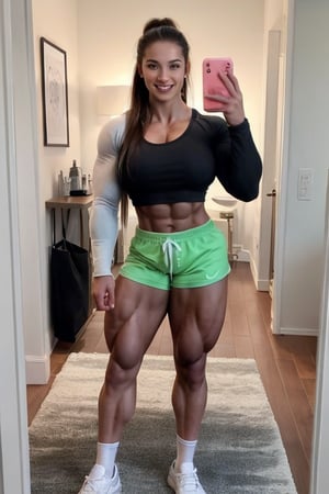 A heavily muscled iffb pro female bodybuilder,  Full-body mirror selfie of a muscular woman standing in a room with a wooden floor. She has long, dark hair with pink highlights and is wearing a black long-sleeve top and light green shorts. She is holding a smartphone with a reflective case in her left hand. She is also wearing white and gray athletic shoes with white socks. The room has a white wall with a calendar or planner hanging on it, and there is a black and white patterned rug on the floor. In the background, there is a black bag and some other items partially visible.