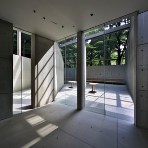 “An interior view of Tadao Ando’s Sendai Mediatheque transformed into an exhibition space focused on architectural and interior design. The gallery showcases minimalist structures and modernist furniture pieces that echo Ando’s signature use of concrete, light, and space. The atmosphere is serene and contemplative, with natural light filtering through the large glass facades, casting soft shadows on the polished concrete floors. The exhibition layout is clean and organized, with each display harmonizing with the surrounding architectural elements. The interplay of light, shadow, and space creates a dynamic yet tranquil environment that invites visitors to engage with the designs on a deeper level.”