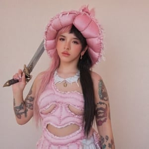 a girl who is caressing the head of a large, pastel pink dragon, while she carries a sword in her right hand while her left hand scratches and caresses the dragon's head, light pink dragon, the girl has fluffy pink and black hair black split dye , 4k, 18k, hairstyle, perfect face, After School, Melanie, Adele, Long hair, masterpiece high quality 18K, Hyperrealism, Realistic vision, rosy cheeks, realistic woman, a girl, Woman, best quality, woman, high quality, good lighting, A woman, with split hair, After School, girl, beauty, a girl who is petting a large light pink dragon with wings, she has her left hand caressing and scratching the dragon's head while her right hand holds a sword, she has fluffy pink and black hair, black split dye, pastel pink dress and hat, dragon, dragon sacrifice, sword, dragon's blood, sword, petting dragon's head, open book ep, tattoos on her arms, dark brow eyes, pink lips, straight hair