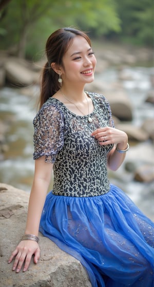 a woman, seated on a rock, wearing a black and white printed dress, adorned with a blue skirt. Her hair is pulled back in a ponytail, and she is smiling. She is wearing a silver bracelet, and a silver necklace. The woman's left hand is resting on the rock, while her right hand rests on her hip. The background is blurred, suggesting a natural setting, with a stream of water and trees in the background.