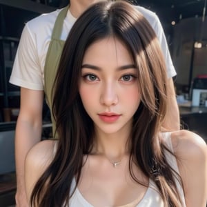 a close-up shot of a young Asian woman with long, wavy brown hair and dark eyes, is positioned in a seductive pose. She is wearing a silver necklace with a pendant, adorned with a silver charm, adding a touch of charm to her outfit. The backdrop is blurred, revealing a man in a white t-shirt and a green apron. The man's arm is positioned behind her, adding to the overall composition.