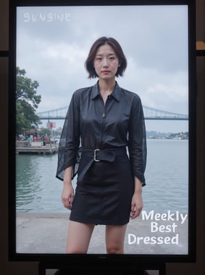 Captured on a digital screen, a woman stands in front of a body of water. She is dressed in a black leather jacket, a black skirt, and a black belt. Her hair is styled in a sleek bob, and her eyes are focused on the camera. The woman's hair is dark brown, and she is wearing a pair of black laces. The backdrop is a cloudy sky, and there is a bridge in the distance. To the left of the woman is a tree with green leaves, and the word "SUNSHINE" is visible in the top left corner of the screen. Below the screen, the words "Weekly Best Dressed" are written in white.