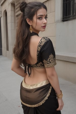 The model is wearing a black saree with a contrasting pink blouse. The saree features a gold border, and the blouse has a halter neck with a back tie and tassels. She accessorizes with statement earrings and a bindi on her forehead. Her hair is styled in a loose, wavy ponytail. The overall look is elegant and contemporary, highlighting traditional attire with a modern twist. The setting appears to be an urban environment with neutral tones in the background, making the colorful saree stand out.