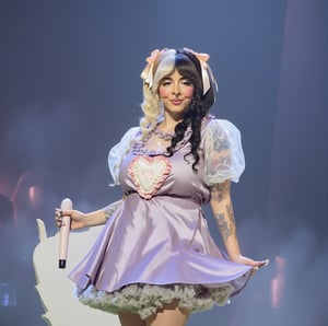 Melanie Martinez stands proudly on stage, her iconic ponytails styled with split hair featuring shades of blonde and black. She wears a striking light purple dress, adorned with bows that add to her whimsical charm. The light purple dress underneath is a nod to her trilogy tour's K-12 theme, with the phrase embroidered on her chest on a giant white patch. The embroidered words "K-12" are embroidered with a light pink thread. Her flawless face shines under the spotlight, capturing the essence of perfection. The full-body shot showcases her statuesque figure, as if frozen in time amidst the enchanting atmosphere.