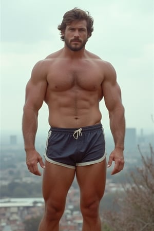 A full-length photograph of a mid-30s man in the 1970s. He is wearing short athletic shorts and no shirt. His beefy, well-worked body stands out. Hair on his armpits, legs, chest and arms. The city can be seen in the background.