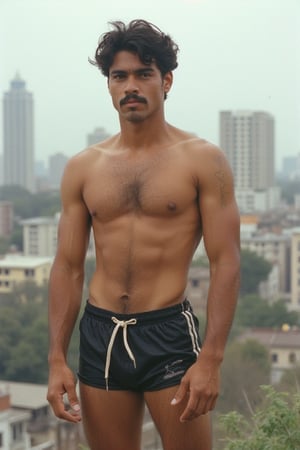 A full-length photograph of a mid-20s latino young man in the 1980s. He is wearing short athletic shorts and no shirt. He has a small moustache. His beefy, well-worked body stands out. Hair on his armpits, legs, chest and arms. The city can be seen in the background. Retro film, noise film, Kodak 400.