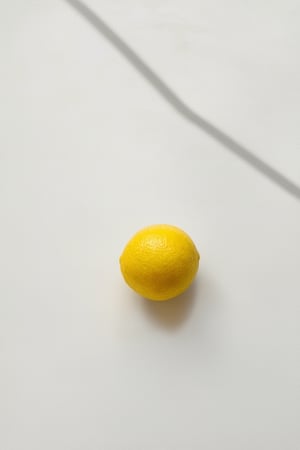 A minimalist still life setup featuring a single yellow object, such as a lemon or banana, placed on a clean white surface. The yellow object is centered in the frame, with plenty of negative space around it to emphasize its bright, cheerful color. Soft natural light illuminates the scene from above, casting a warm glow over the entire composition.