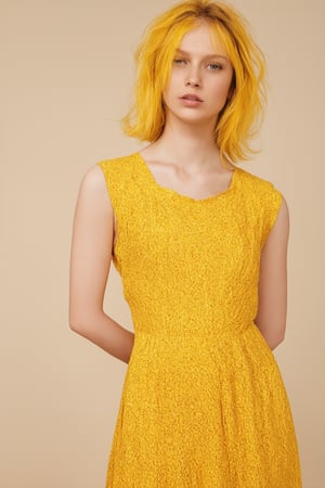 A close-up shot of a retro-style color model, 'Basic Yellow', set against a warm beige background. Soft focus and gentle lighting accentuate the model's bright yellow hair and matching dress, with subtle highlights on the fabric and a slight sheen to evoke a sense of nostalgia. The subject stands relaxed, hands behind her back, exuding confidence and playfulness, as if about to burst into a sunny day.