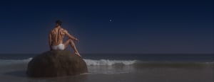 Photorealistic 8k RAW photo of a full-body muscular man with a handsome face and tan skin, sporting short black hair and a clean-shaven face. He is posed elegantly on a large moonlit boulder by the sea, with gentle waves lapping at his feet as he gazes out at the starry night sky. The image is ultra-detailed with high-resolution clarity, natural lighting, and a focused composition on his powerful pose and the serene, moonlit setting.