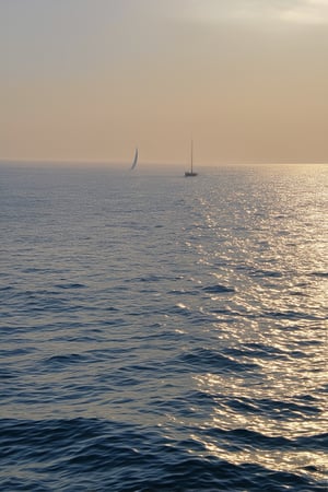 A serene seascape: a misty dawn breaks over the tranquil waves of DeepBlue, where iridescent mist swirls, veiling the horizon. The ethereal atmosphere is set by soft, golden light casting an otherworldly glow on the rippled water's surface. A lone sailboat drifts, its white sails billowing in the gentle breeze as it disappears into the shrouded distance.