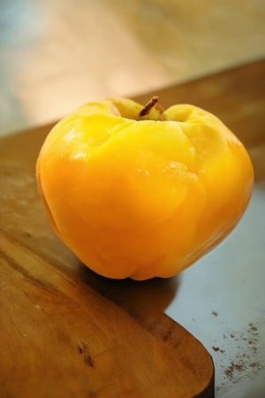 A serene still life featuring a vibrant yellow Hakame (Japanese apricot) prominently centered on a rustic wooden cutting board. Soft golden light falls from above, casting a warm glow on the fruit's smooth surface. The camera frames the subject in a shallow depth of field, blurring the background to emphasize the fruit's delicate texture and subtle sheen.